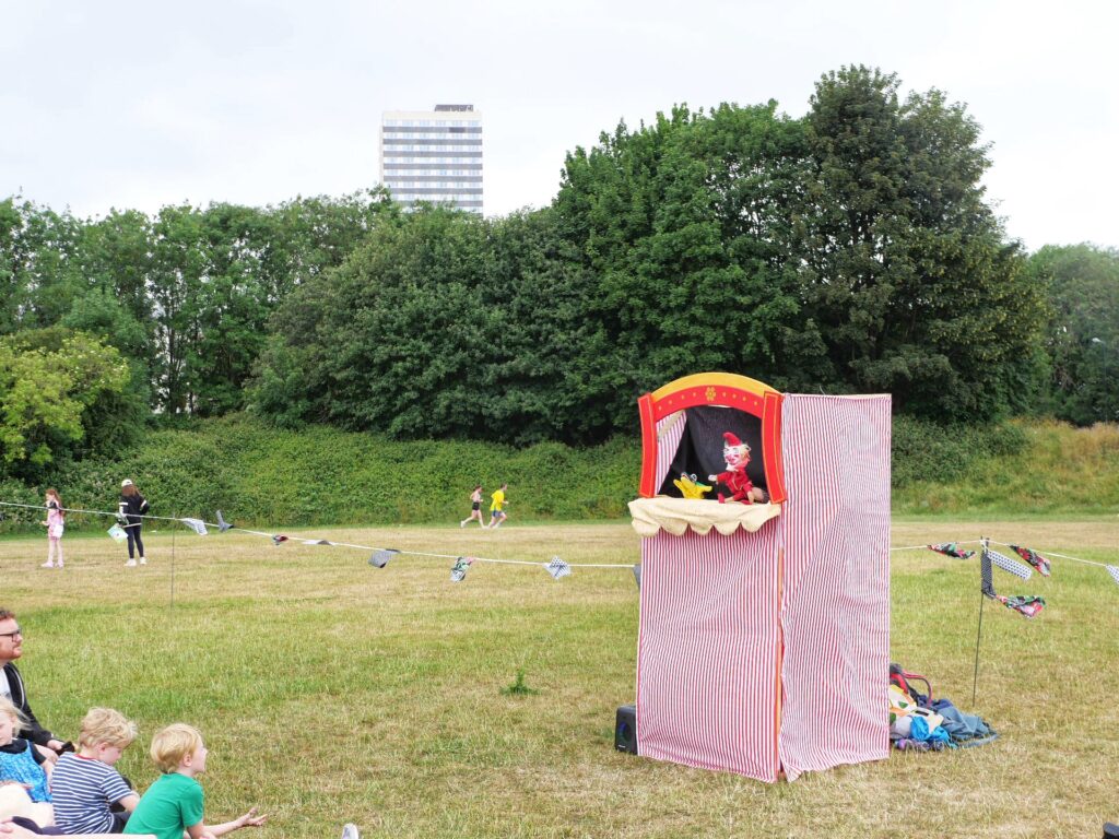 A Punch and Judy theatre on a playing field. In the background, a row of trees, and behind that, a block of flats.