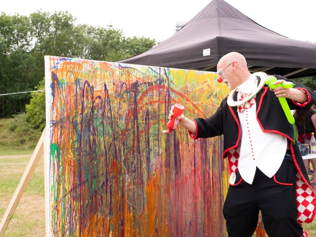 A clown wearing a black, red and white circus outfit squirts red paint at a messy canvas