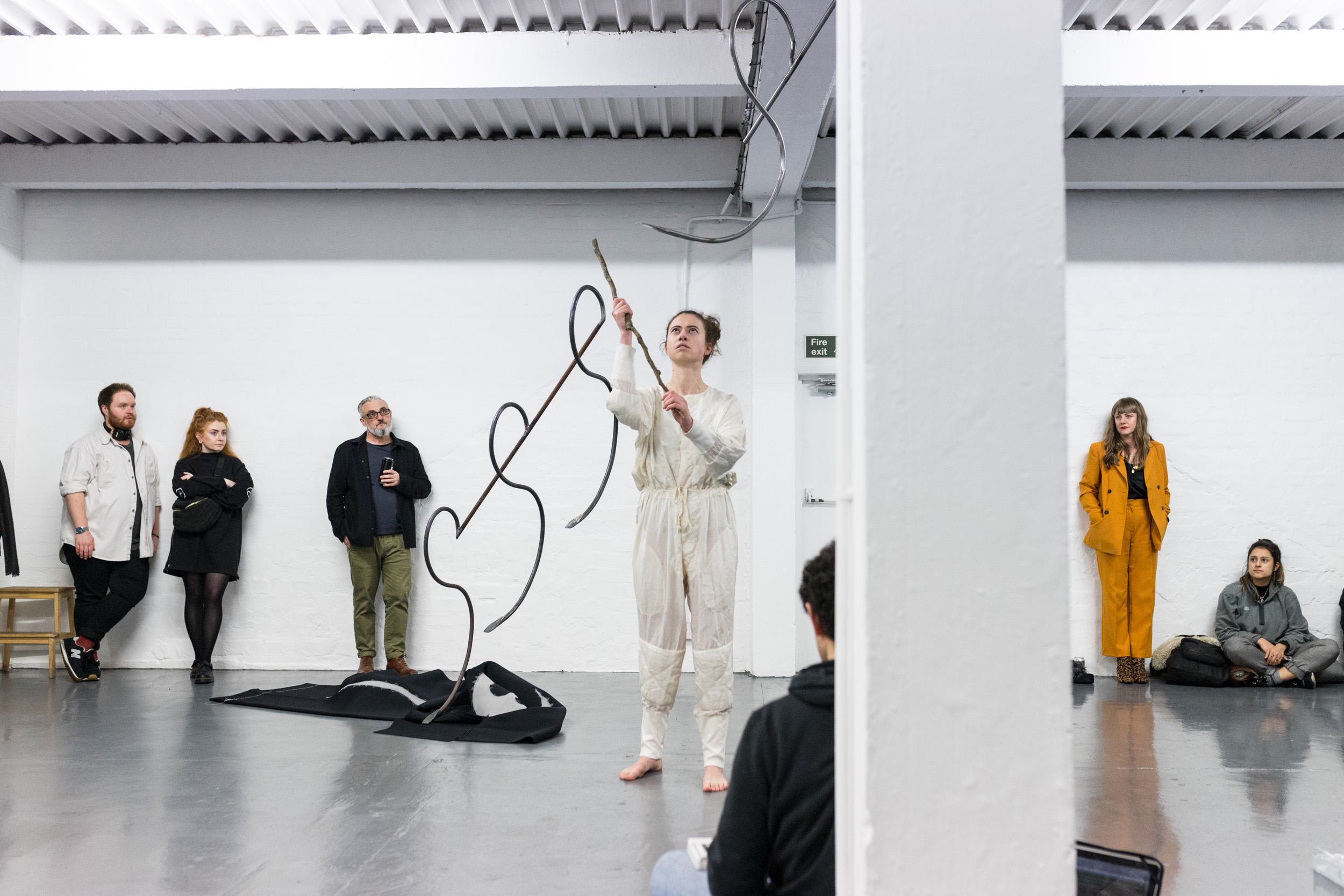 Installation view of a performance at Grand Union featuring a female dressed in white interacting with tangled metal objects with an audience in the background
