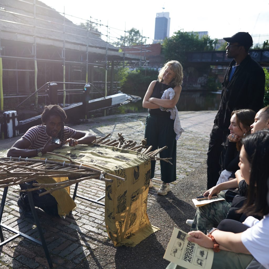 An outside scene and a group of people watch a man (part of RESOLVE Collective) talking whilst kneeling down twining twigs and fabric together