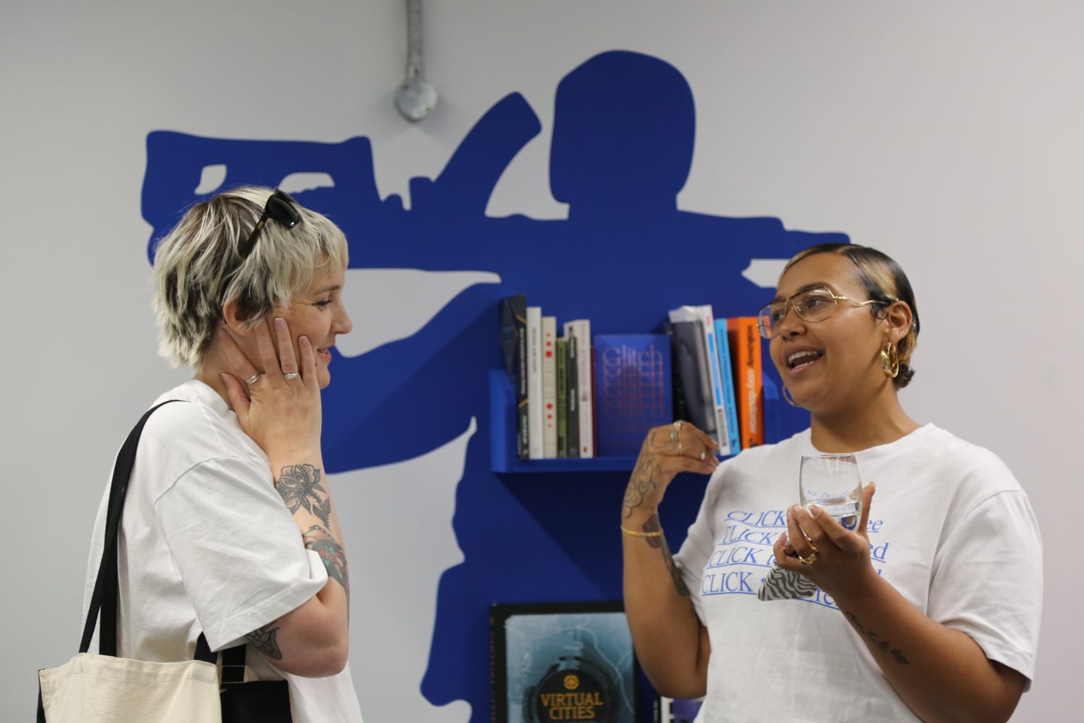 Two women speak together. One woman is light skinned the other is dark skinned. Both wear white t-shirts. Behind is a bookcase whose silhouette resemles a soldier carrying a gun.