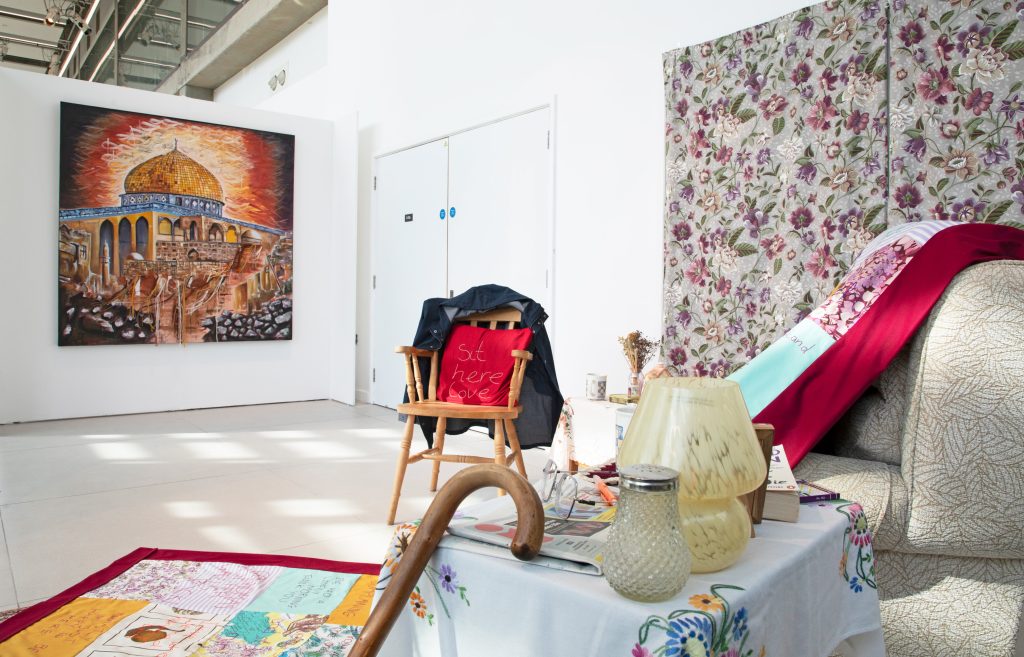 A large square painting on the back wall of a white-walled gallery. In the foreground are some chairs and domestic items that look as if they have been lifted directly out of a scene in sombody's home.