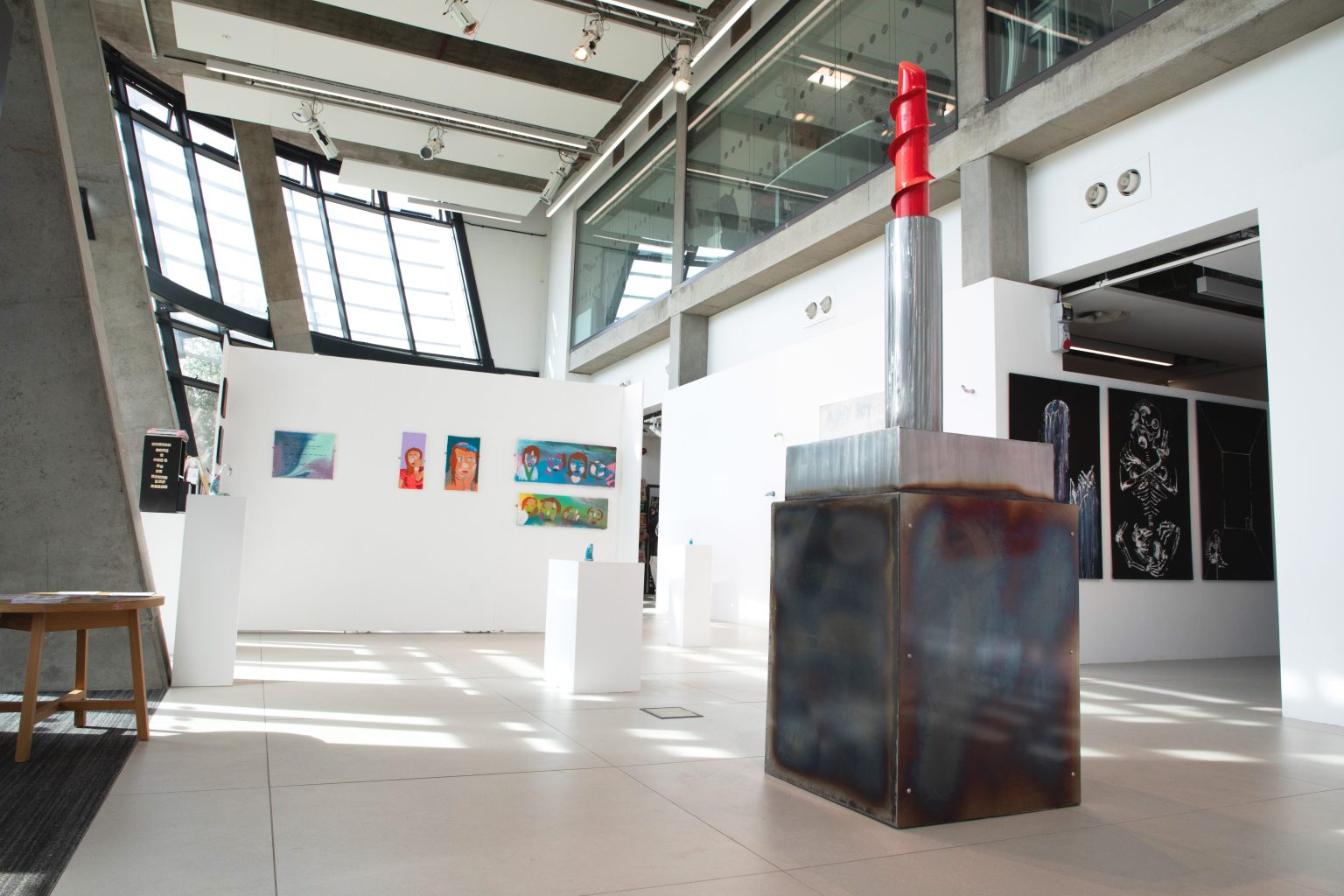A double height gallery space with white walls and concrete pillars containing a range of different artworks in a variety of media and sizes.