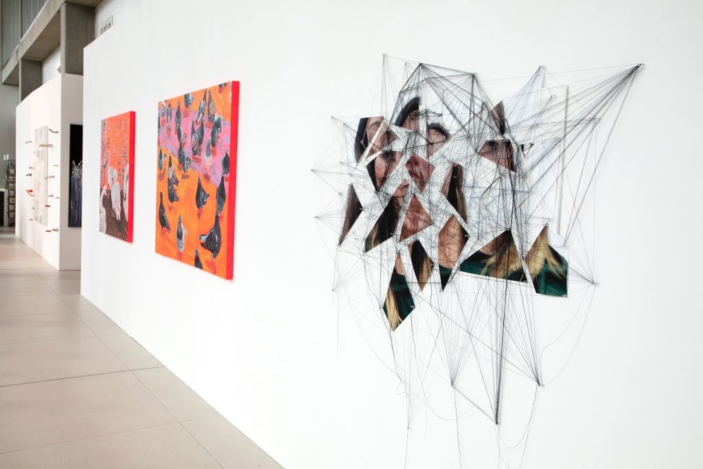 Two brightly coloured orange and red paintings sit on the left of a long white wall. On the right and foregrounded is an artwork that is comprised of fragmented images - parts of a face perhaps - connected with black threads.