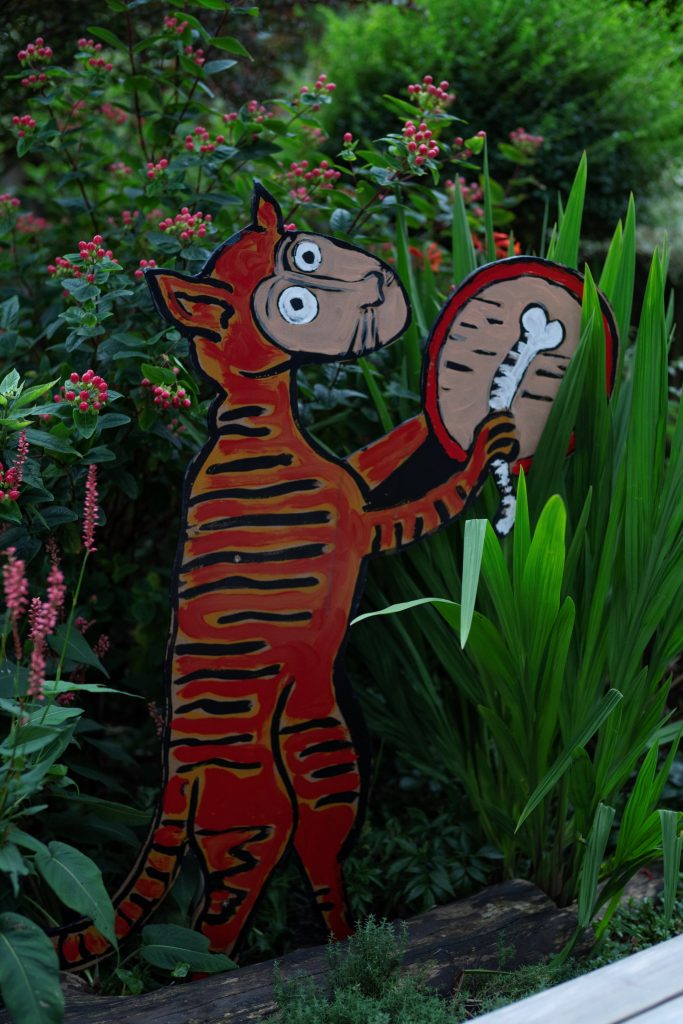 A painting of a dog playing a drum with a bone made on dummy board and stood amongst vegetation.