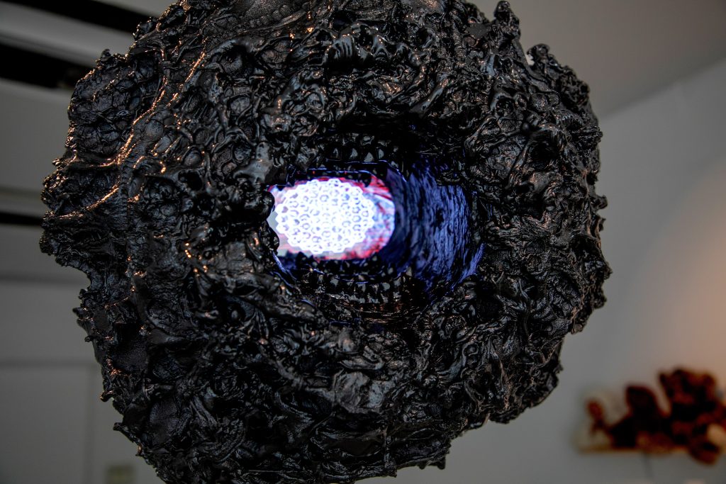A large black sphere with a rough textured surface with a video screen illuminated at the centre. In the background, out of the depth of field, is another sculptural object.
