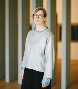 Photo of Elizabeth Price. A light skinned woman with blonde hair wears spectacles, a loose fitted grey jumper and a long black skirt.