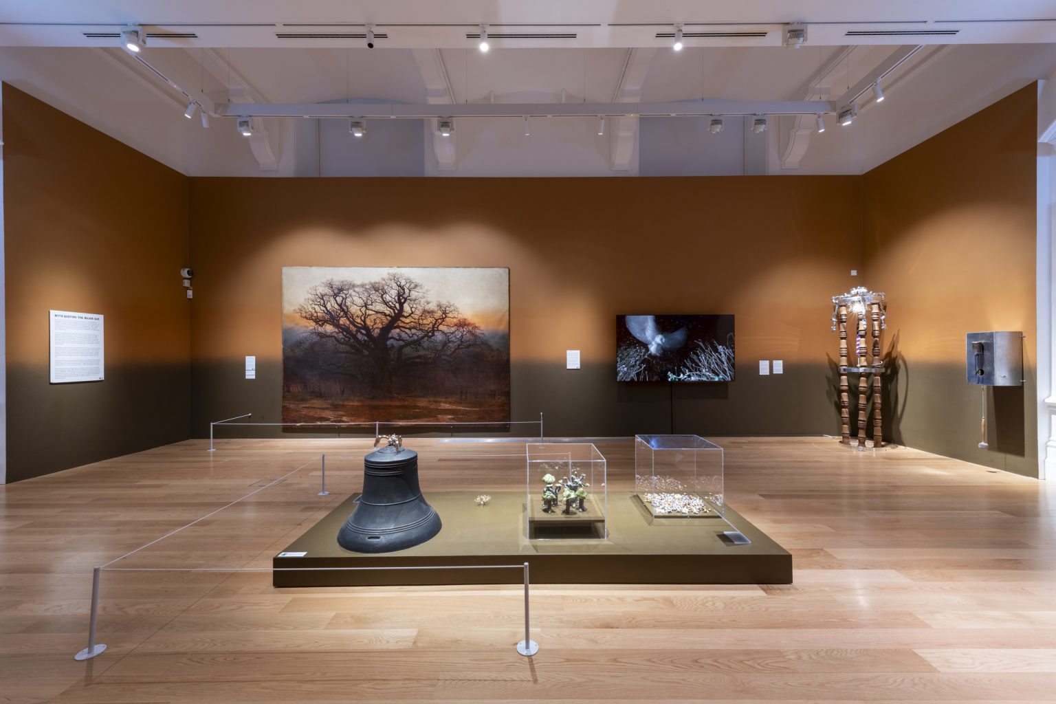 A gallery with walls painted in mid-brown and dark terracotta, with a variety of different works including a large painting, a video piece on a wall mounted screen and several sculptural works in the foreground on a low plinth.