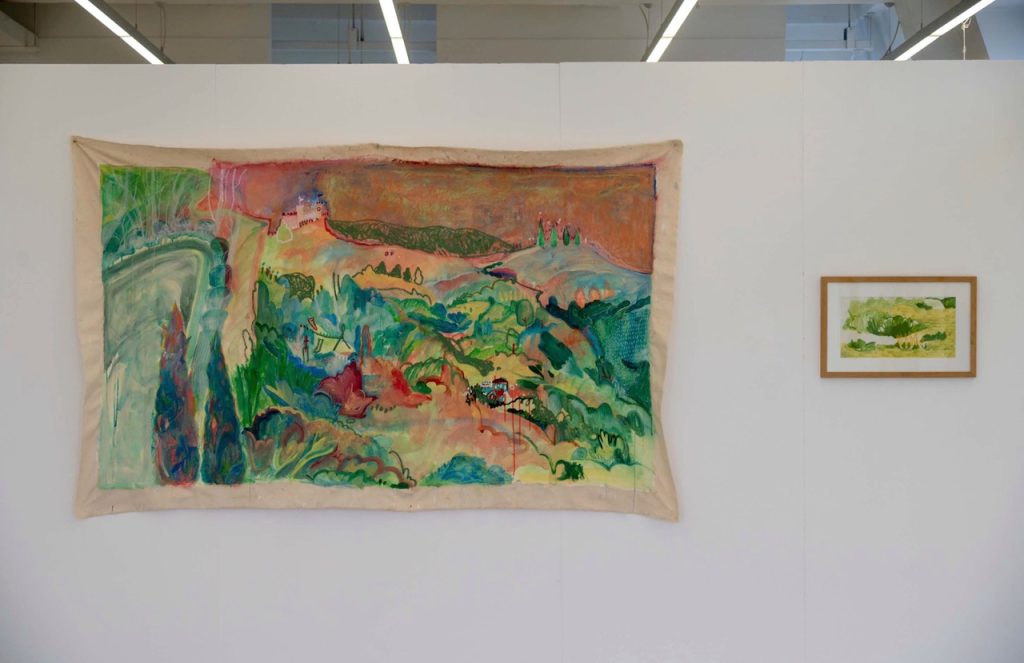 A white gallery wall. On the left a large, unstretched canvas with a colourful landscape painted on, to the right a smaller painting, a detail of grass in yellow and green, in a small brown frame.