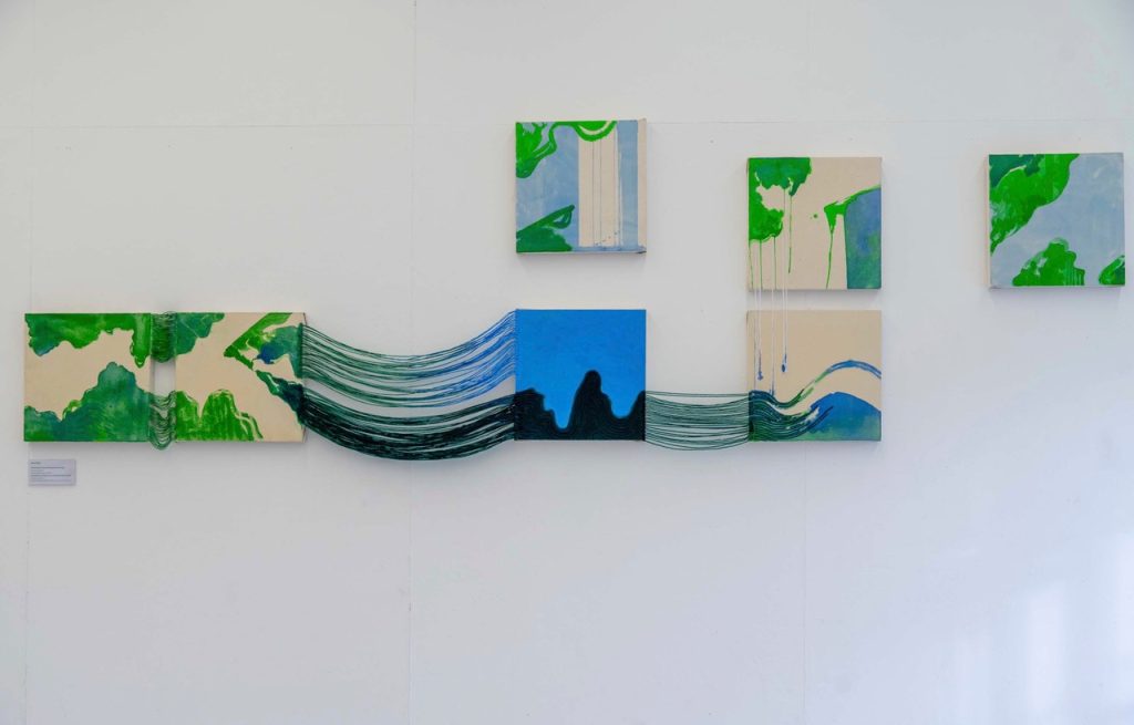 A white gallery wall. Seven small square panels, painted in semi abstract blue, green and black forms. Four of the panels are connected by coloured strings suggesting the paint is coming off one panel into the next.