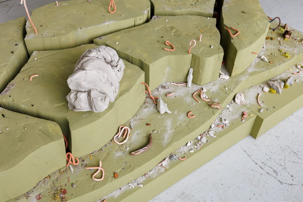 close up of a green moss coloured plinth covered in pink clay worms and grey dust