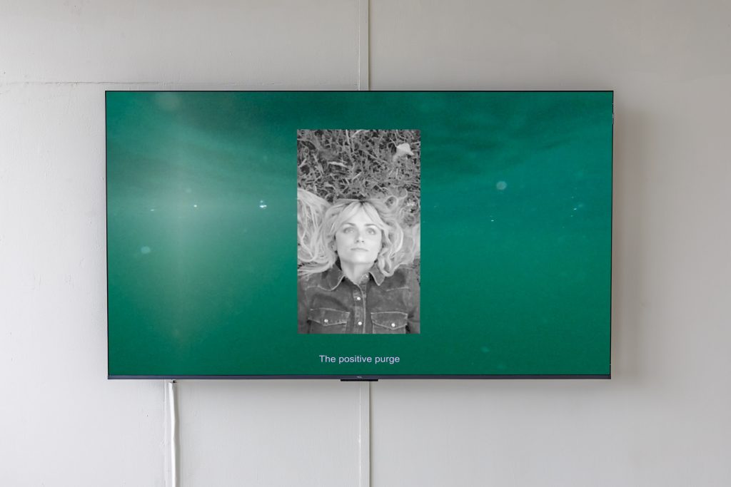 tv monitor showing turquoise water and a greyscale portrait of the artist with the words 'the positive purge'