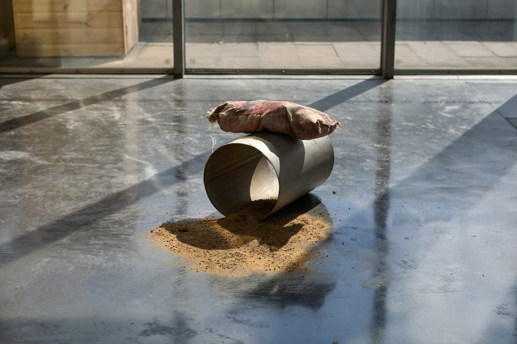 A steel tube the size of a bucket lay on its side atop a pile of sand and gravel and has a sandbag on top