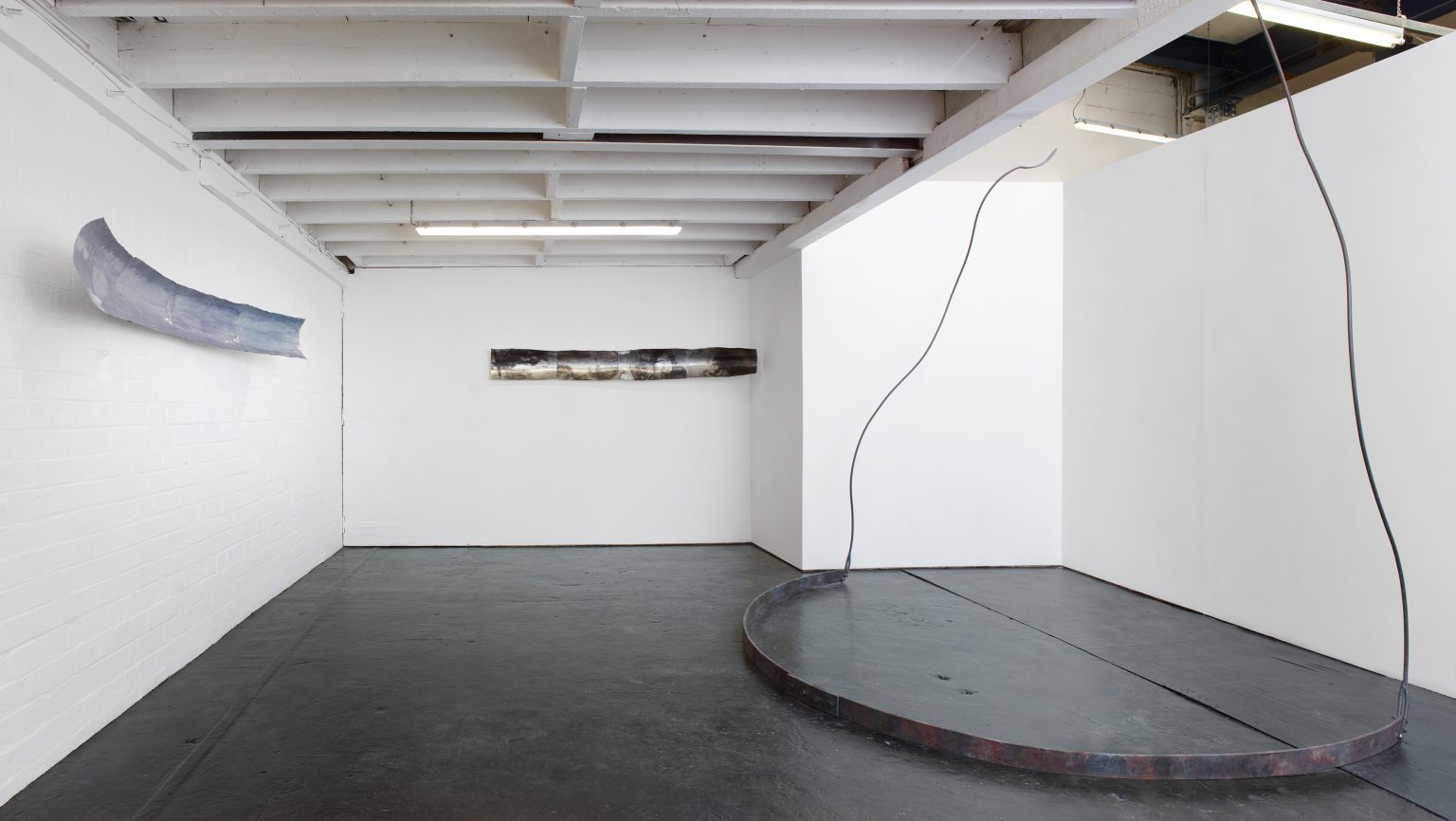 A white wall gallery space with a grey floor. On the floor to the right and in the foreground, a curved piece of steel with two tentacular twists of metal leading from each end up into the air, towards a skylight above. On each of the two back walls, strips of metal at around head height - one light grey in colour, the other with darker markings.