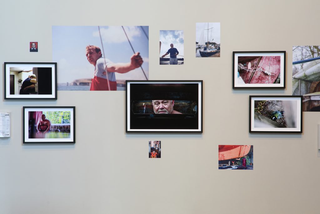 A collection of framed photographs show the artist's father and his boat on the Norfolk Broards - collaged on the wall, some overlapping with images pasted directly onto the wall - all in differing sizes