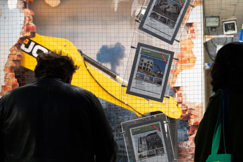 A wire fence with images of properties, like a real estate agents window, but curved. Two viewers stand looking at the images.