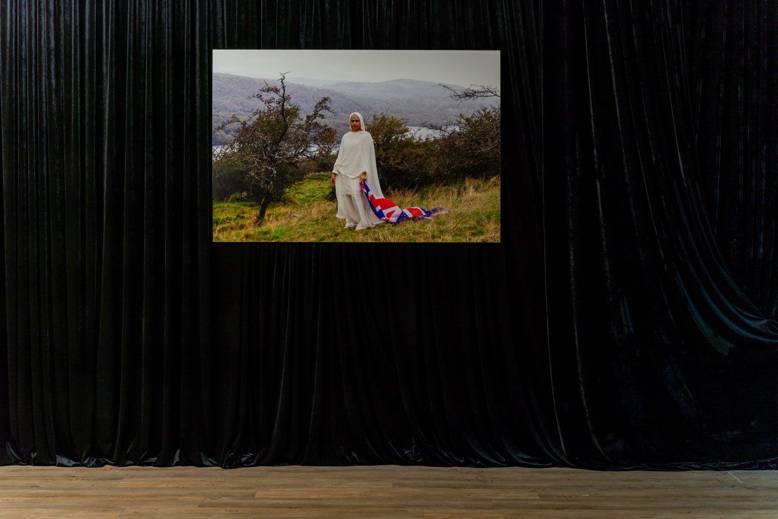 A glossy black curtain with many folds. On it a photograph, large and in colour. It shows a woman with light brown skin standing in a landscape she is wearing a white robe and head scarf, in her left hand a Union Flag hangs, draping on the ground.