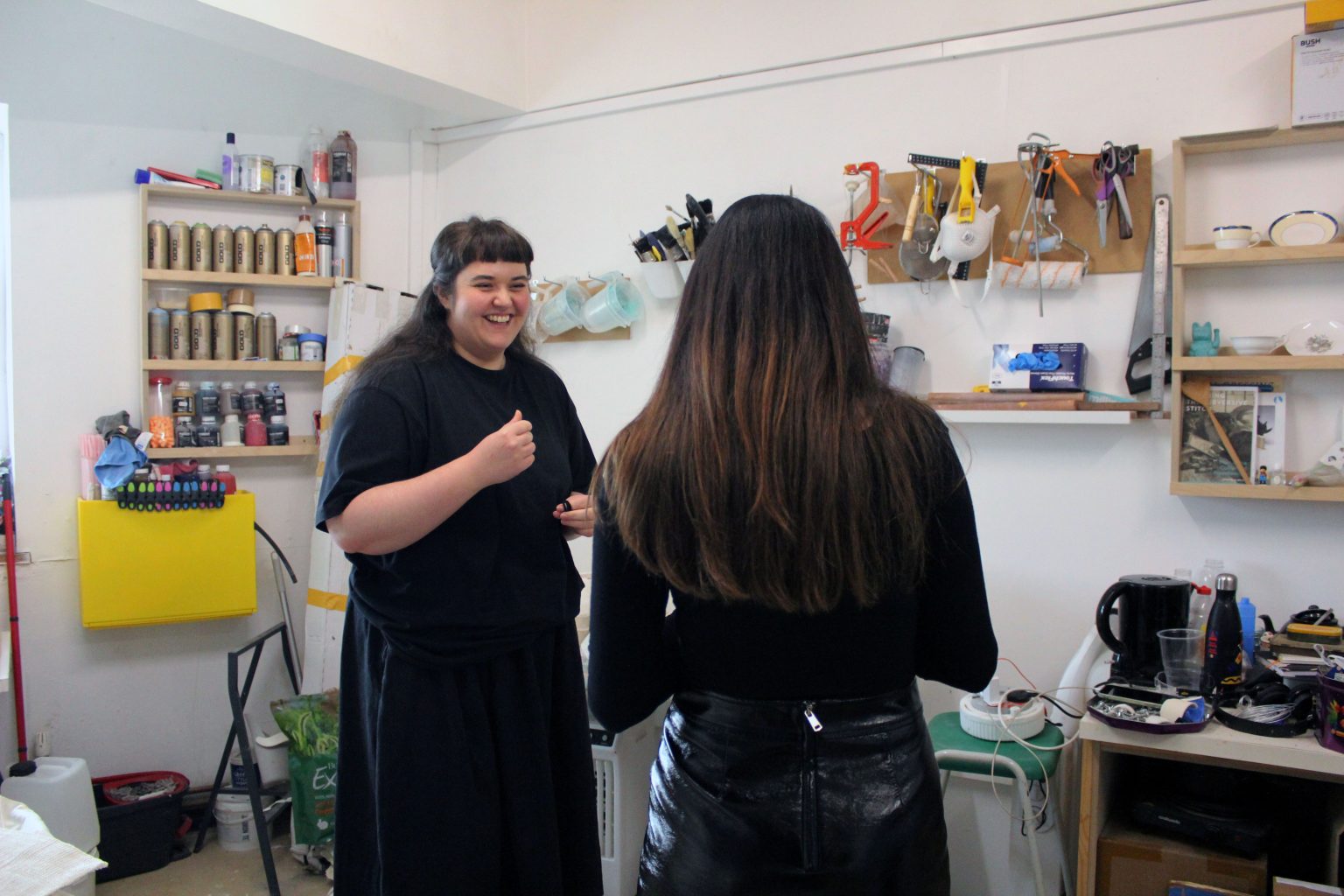Two people in dark clothes with dark, long hair stand chatting and smiling in an artist's studio, surrounded by colourful objects and shelves of artist materials
