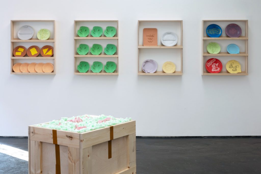 A white wall gallery space. On the floor, a large, unopened packing crate; on the wall behind, four similar shelving units displaying colourful ceramics.
