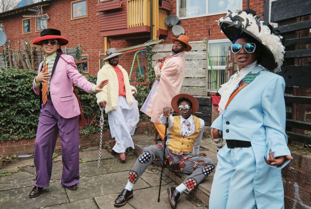 A group of Afro-Caribbean people dressed in bright, colourful and fancy clothes, are posing in the back yard of a council house.