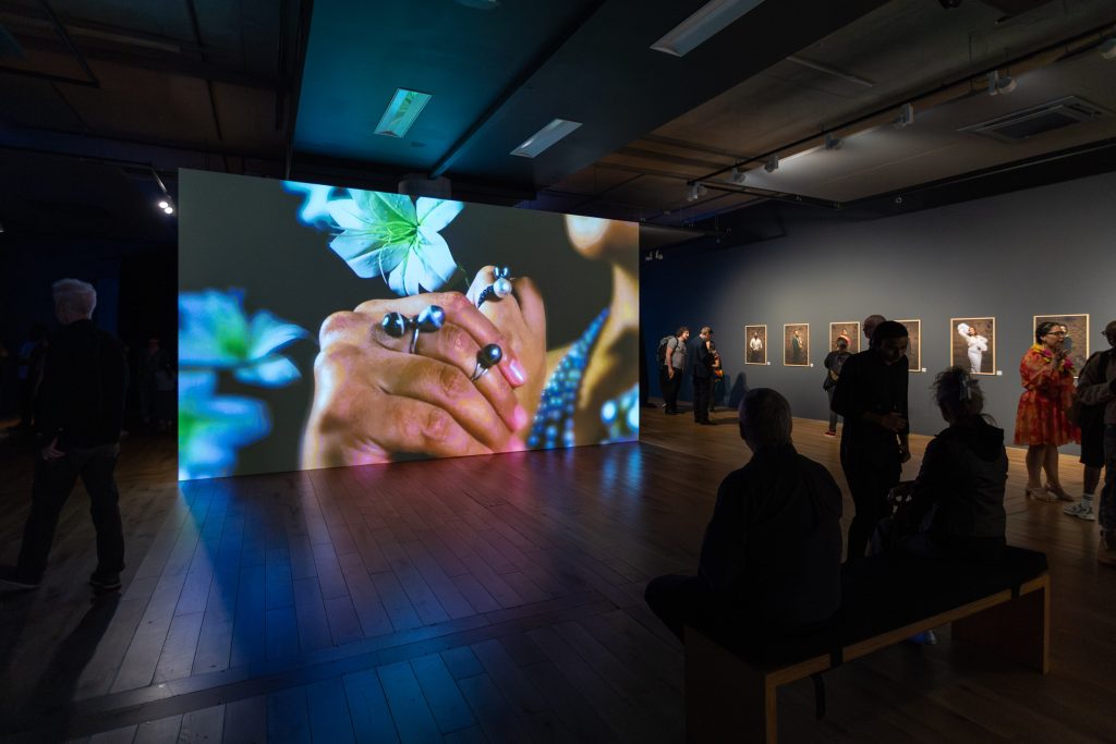 A large screen shows a closeup of clasped hands with rings on the fingers