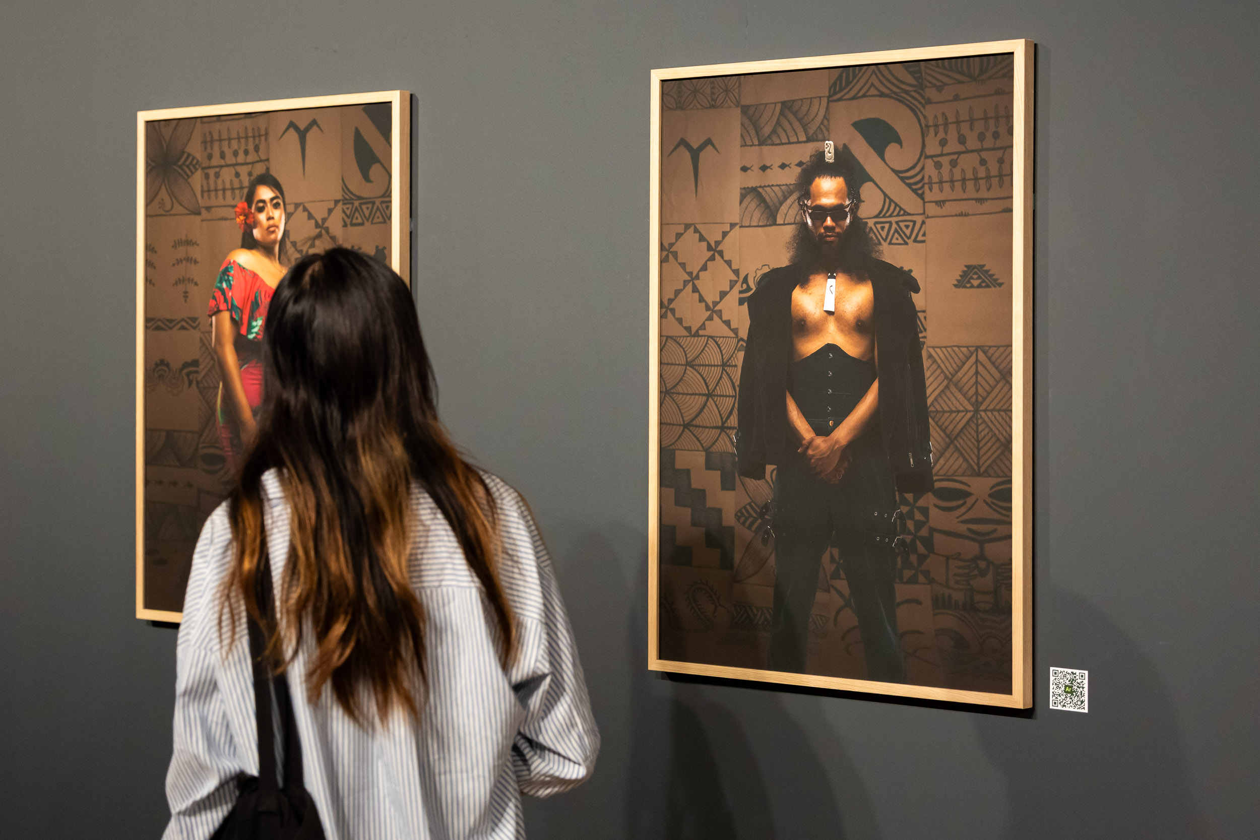 A viewer looks at two framed photographic portraits, on the left a person in a red rdess with a flower in their hair and the right a person in black clothing and glasses