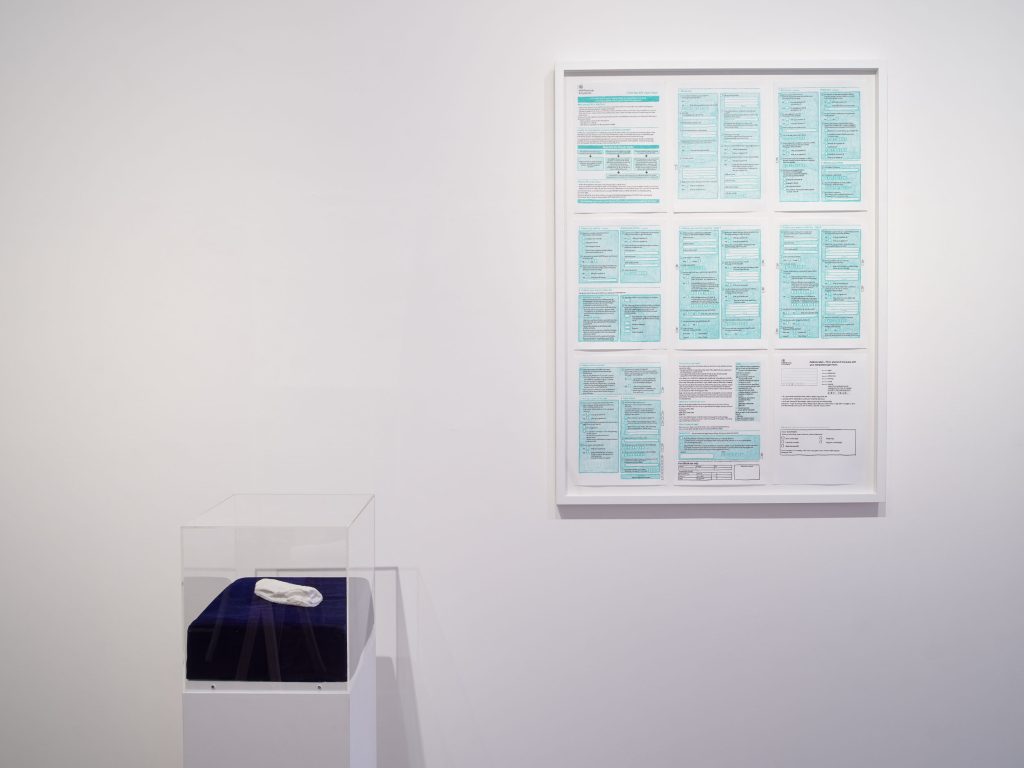 A small vitrine against a white wall containing a small white object. Also on the wall, the framed pages of a benefit claim.
