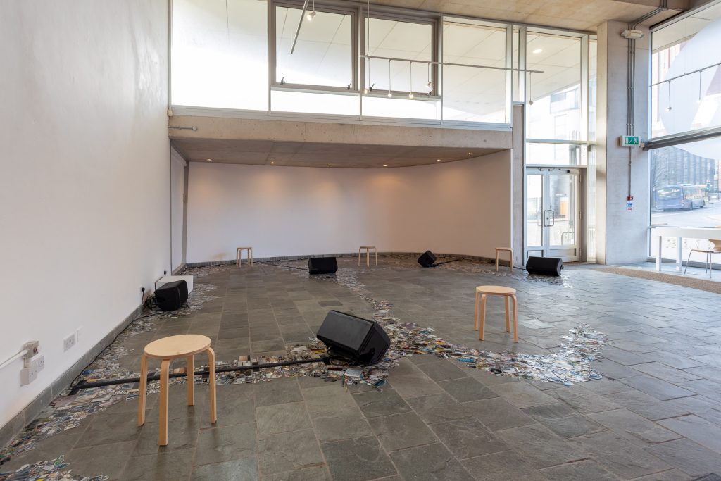 A large white wall gallery space with a tiled floor, across which stretch veins of closely-packed, cut-out images gathered around speaker cables and speaker cables spread around the space.