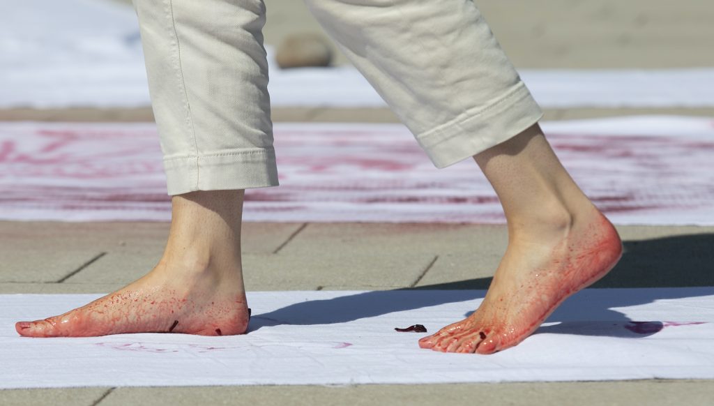 A pair of feet, in profile, in motion walking over a sheet of white fabric. The feet are stained with pink dye.