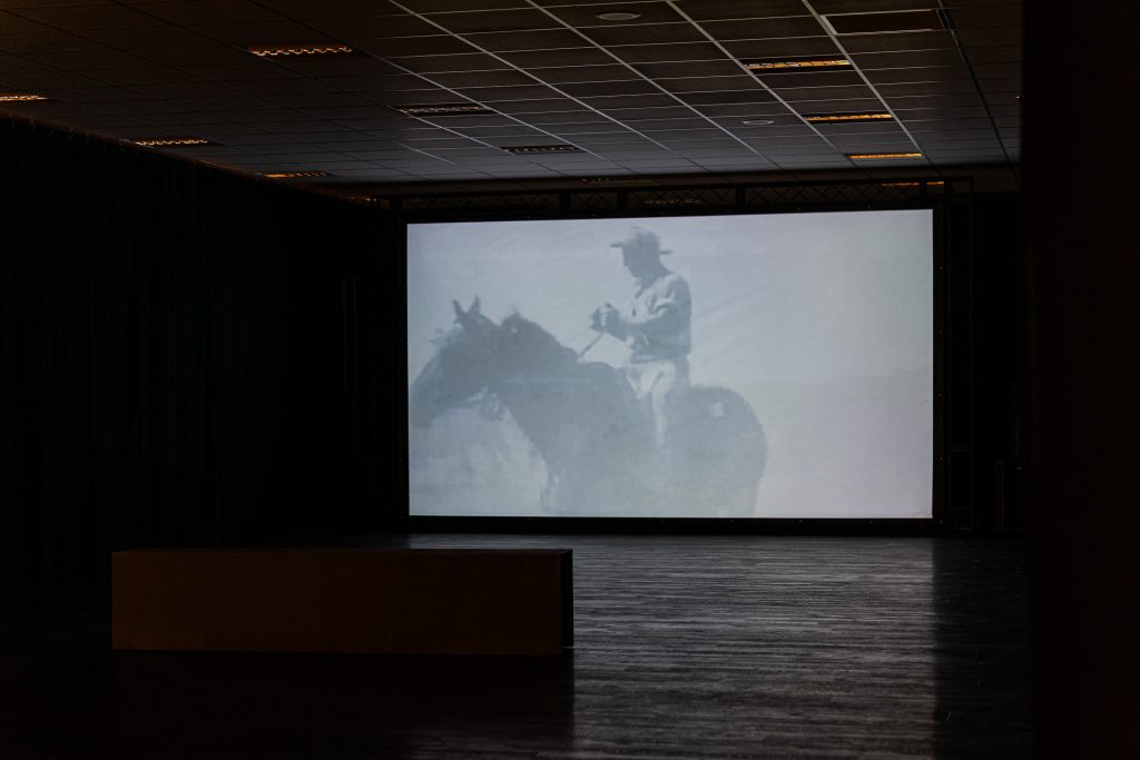 A darkened room with a bench in the middle. Projected onto the far wall is an image of a cowboy on a horse in faded black and white.