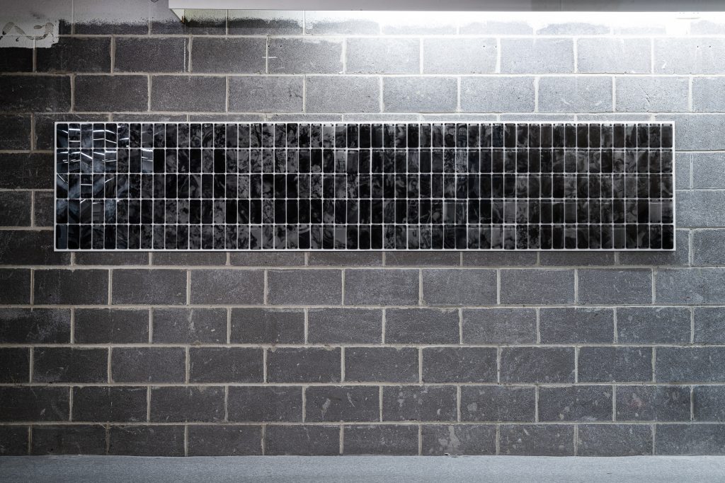 A breeze block wall. On the wall is a mosaic of plastic phone screens, five screens high and approximately sixty screens wide.