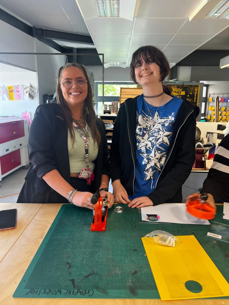 A teacher and student stand at a desk with a cutting board and badge making equipment. They are both smiling.