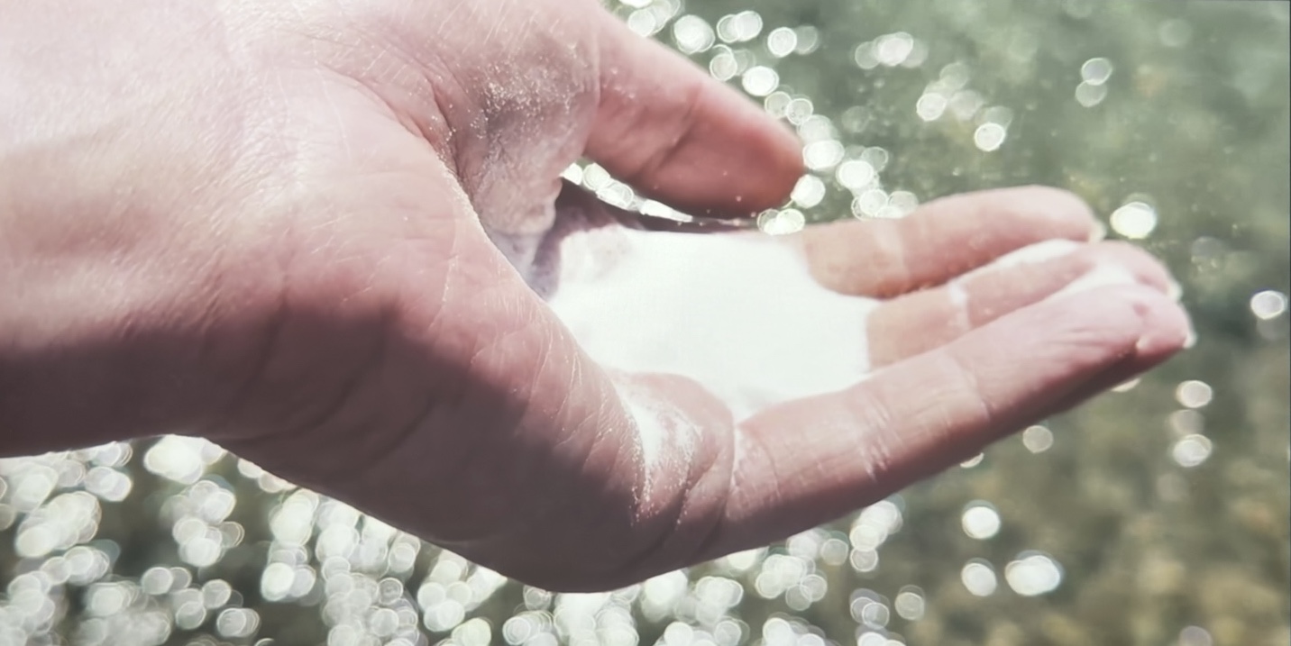 A cupped hand holds enough salt to fill its palm. The hand is held above a glistening body of water.