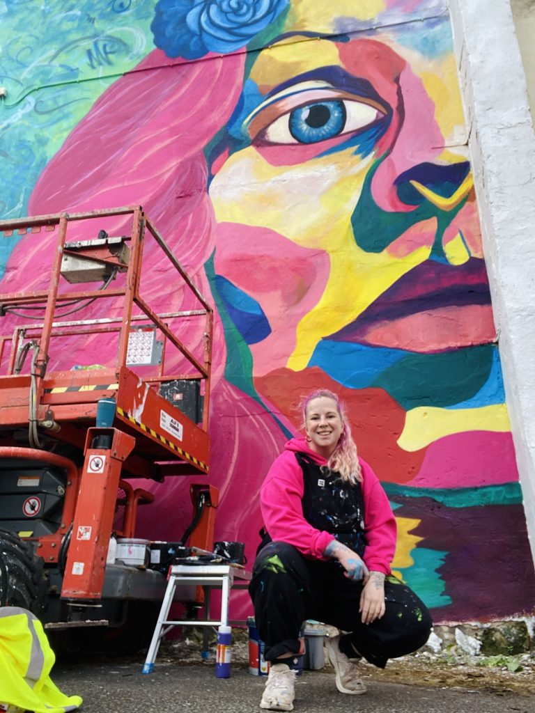 A brightly painted mural of a large face on a wall. A woman poses in front of it kneeling down.