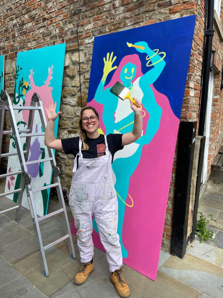 A person in white overalls is holding a paintbrush in the air and smiling widely. Behind her is a large board with a brightly painted figure on it.