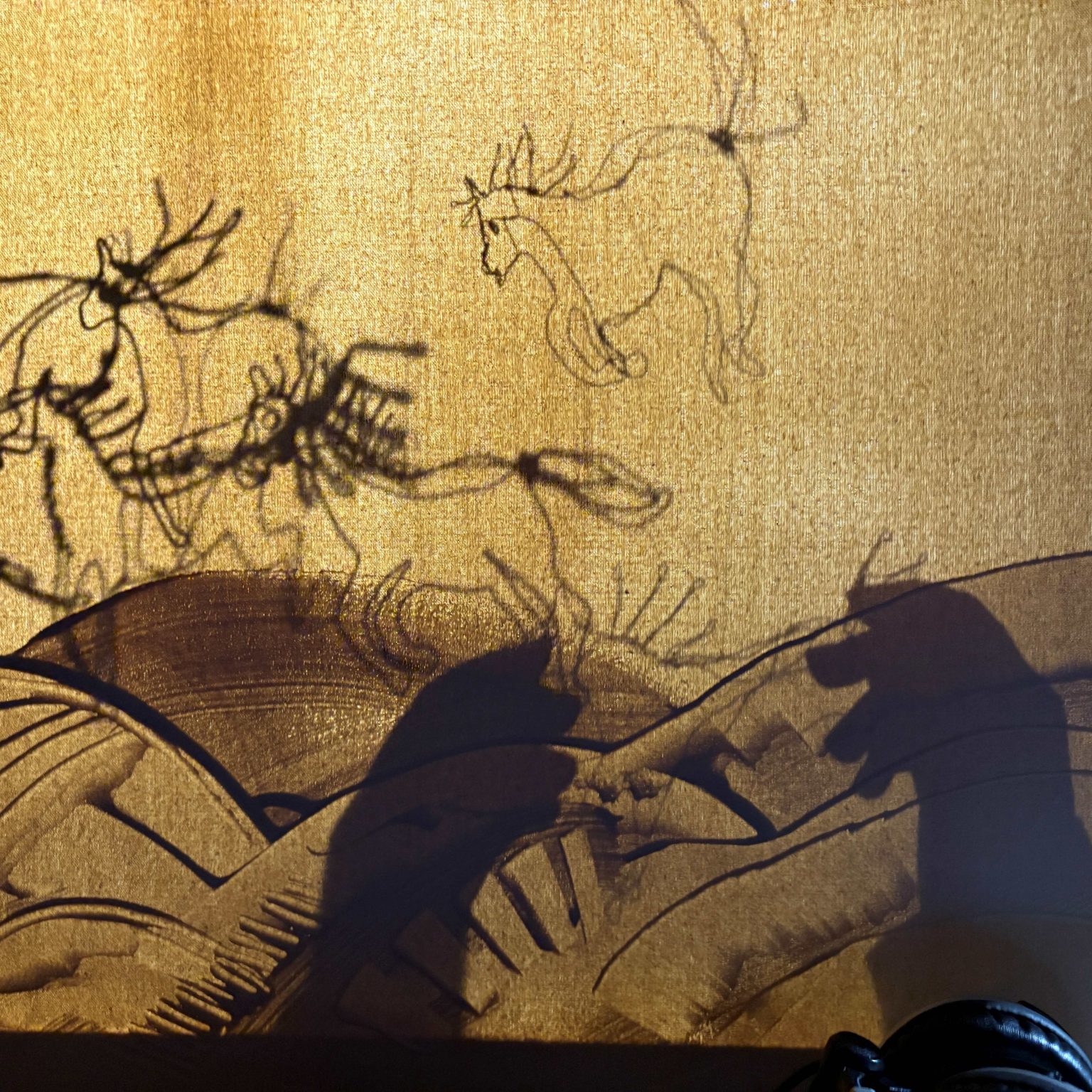 Various shapes cast in shadow on a stretched yellow canvas - illustrative outlines of galloping horses; broad brushstrokes describing hillsides; a pair of human hands.
