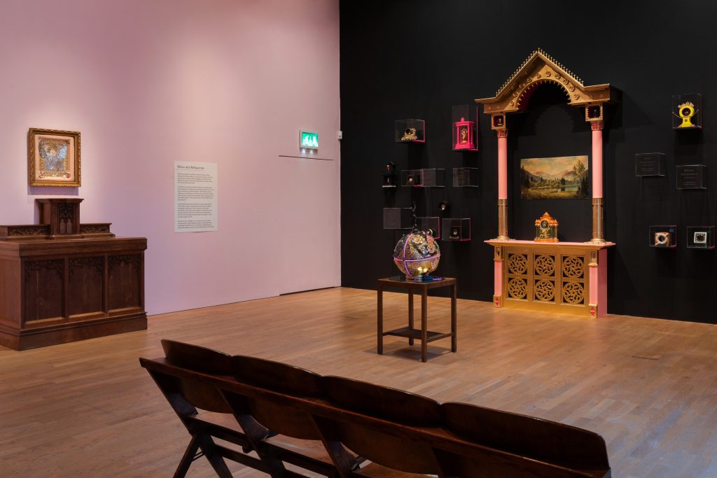 A Gallery with a pink wall and a black wall. On the black wall a kind of shrine with a carriage clock on.