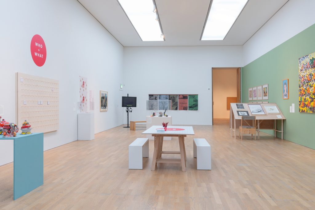 A gallery with white walls and laminate flooring. A table woth benches in the centre of the space. Bright images on the walls. A blue plinth with objects on.