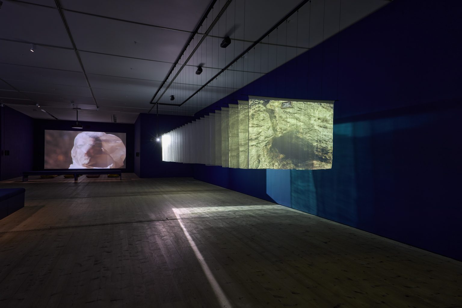 A large gallery. A set of screen-sheets suspended from the ceiling with a bright image projected onto them. To the right, a screen showing a face that seems to be covered in fabric.