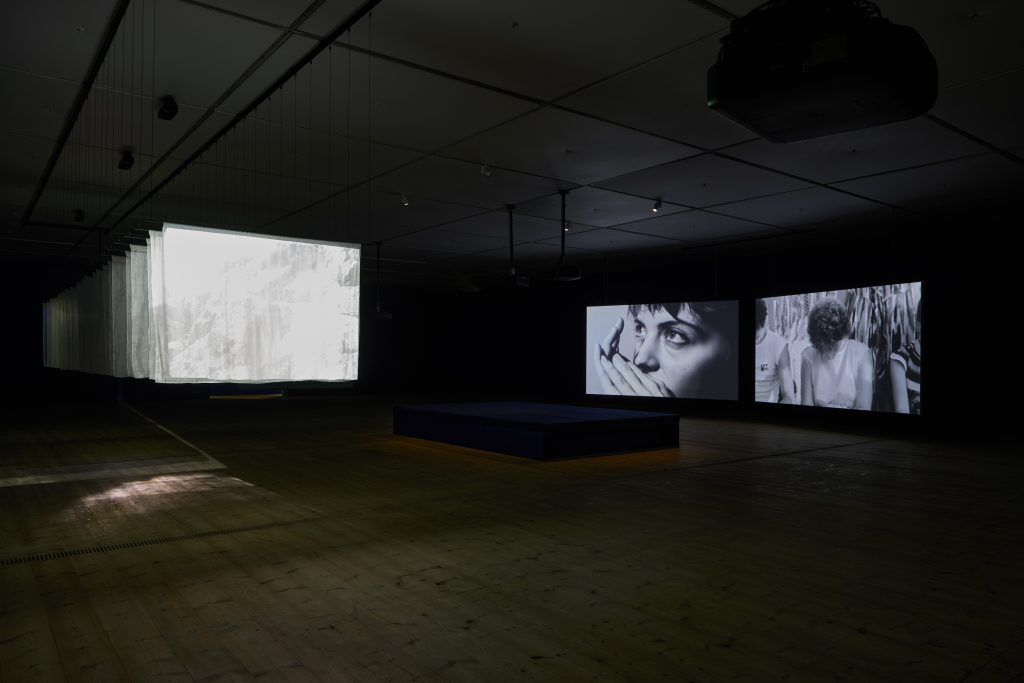 A large gallery. A set of screen-sheets suspended from the ceiling with a bright image projected onto them. To the left, two screens, one with a close up of a woman's face, the other shows three figures sitting, their faces down.