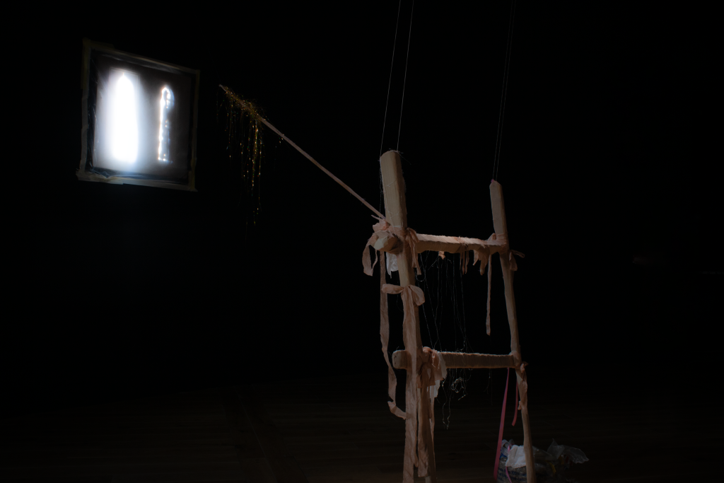 A black image with a dimly light light wooden fence covered with pink ribbons in the foreground, lit by a small, illuminated windowpane above