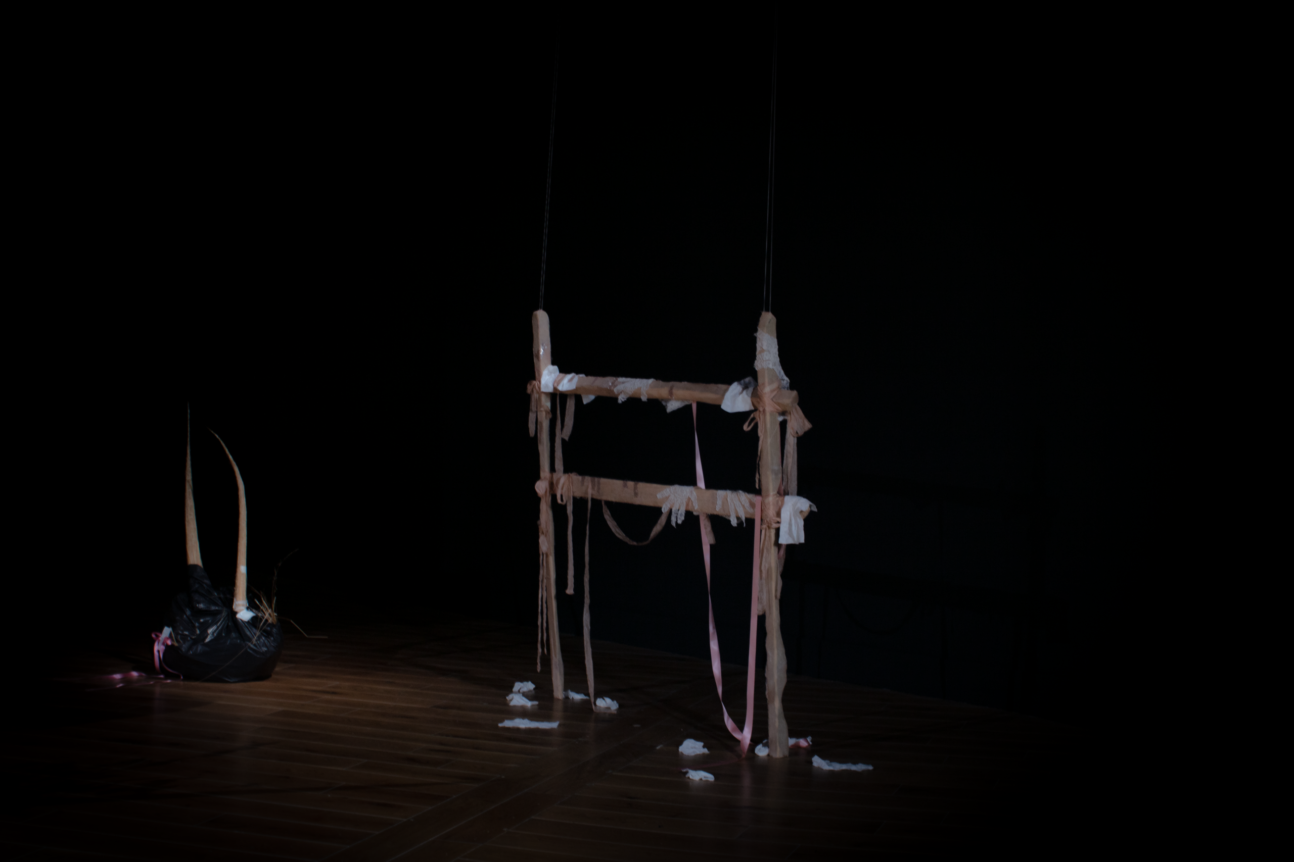 A light wooden frame is dimly illuminated in the darkness, with pink and white ribbons and hand shaped fabrics draped from it