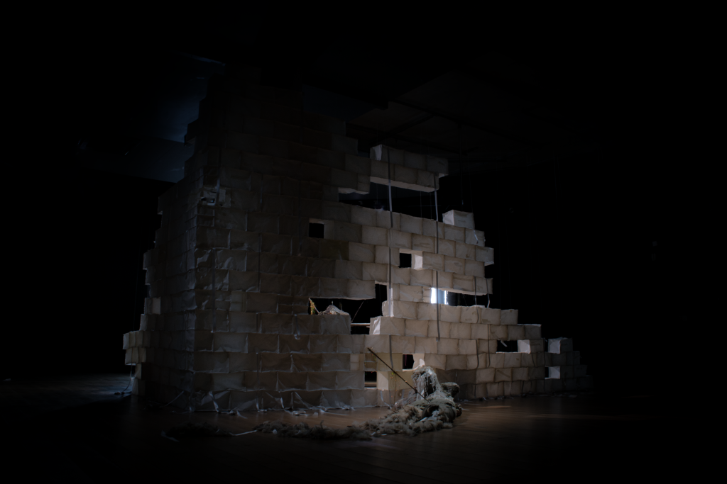 A dimly lit wall surrounded by darkness, built of semitransparent white fabric cubes through which the light shines, illuminating a semi rotted fabric corpse like body in the foreground