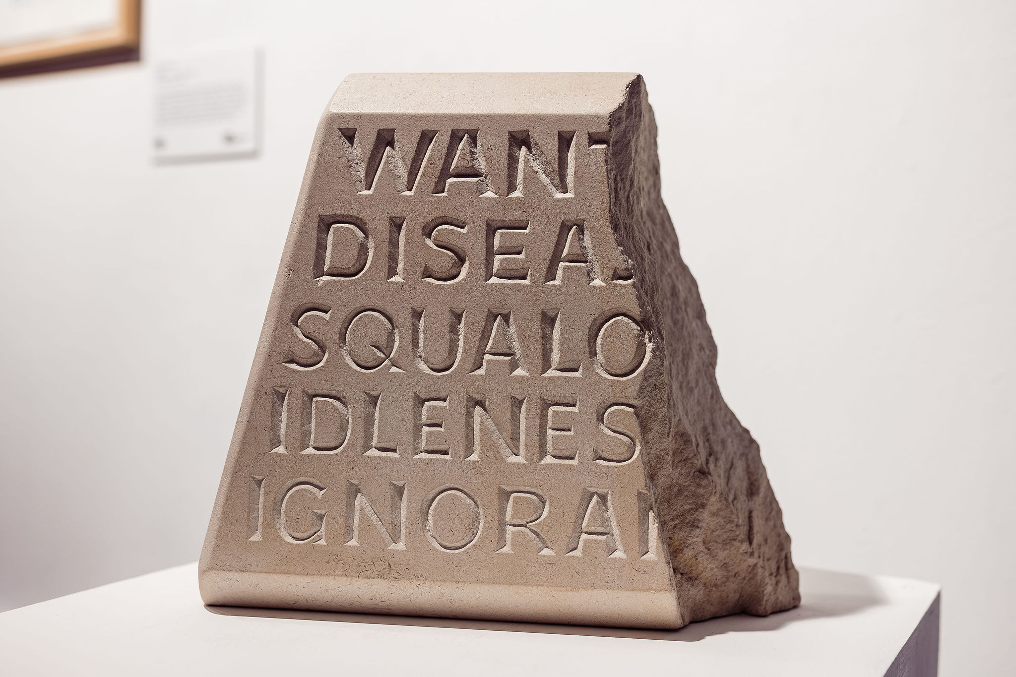 A stone block carved to a smooth shape on the left and rough hewn on the right, interrupting the words carved on it: 'Want-; Disea-; Squalo-; Idlenes-; Ignora-'