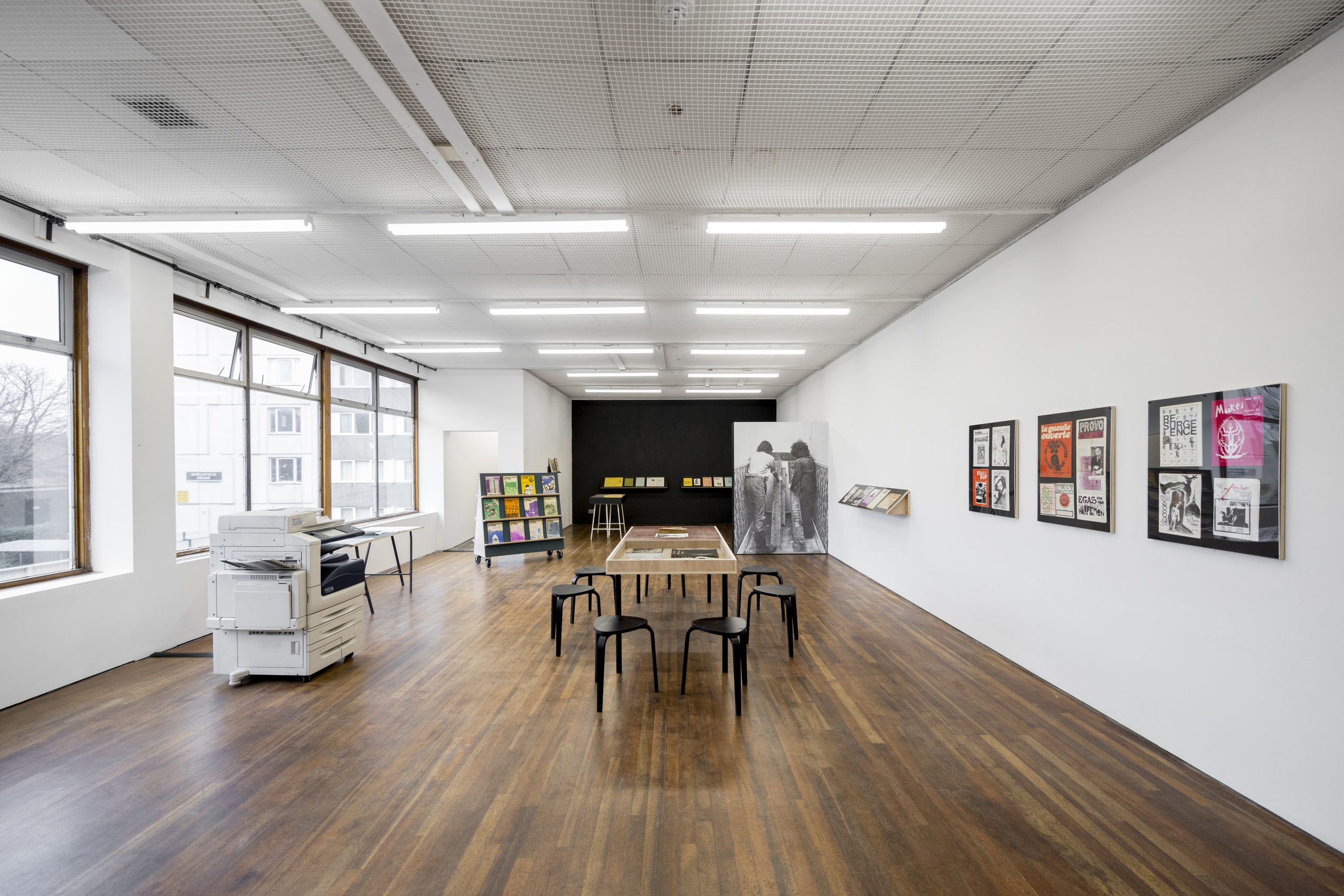 A gallery space with wooden floors and white walls. A large table in the middle surrounded with stools. On the right hand wall three large frames with book covers displayd inside. To the left, large bright windows and a Xerox machine. More images displayed at the back of the gallery.