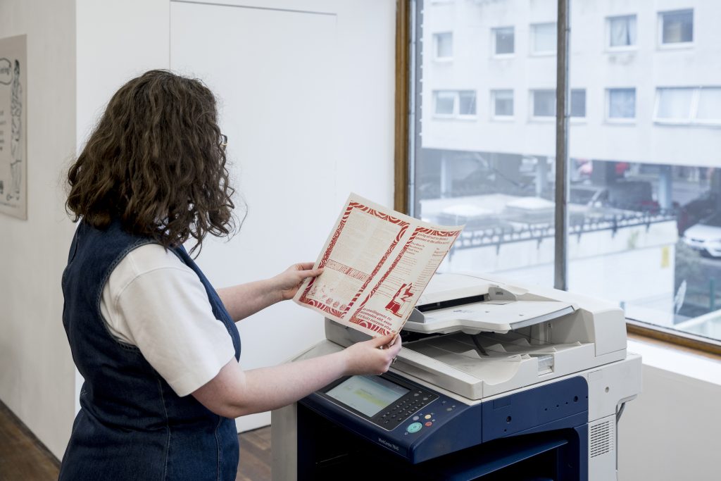 A person with curly hair stands by a Xerox machine holding a pamphlet.