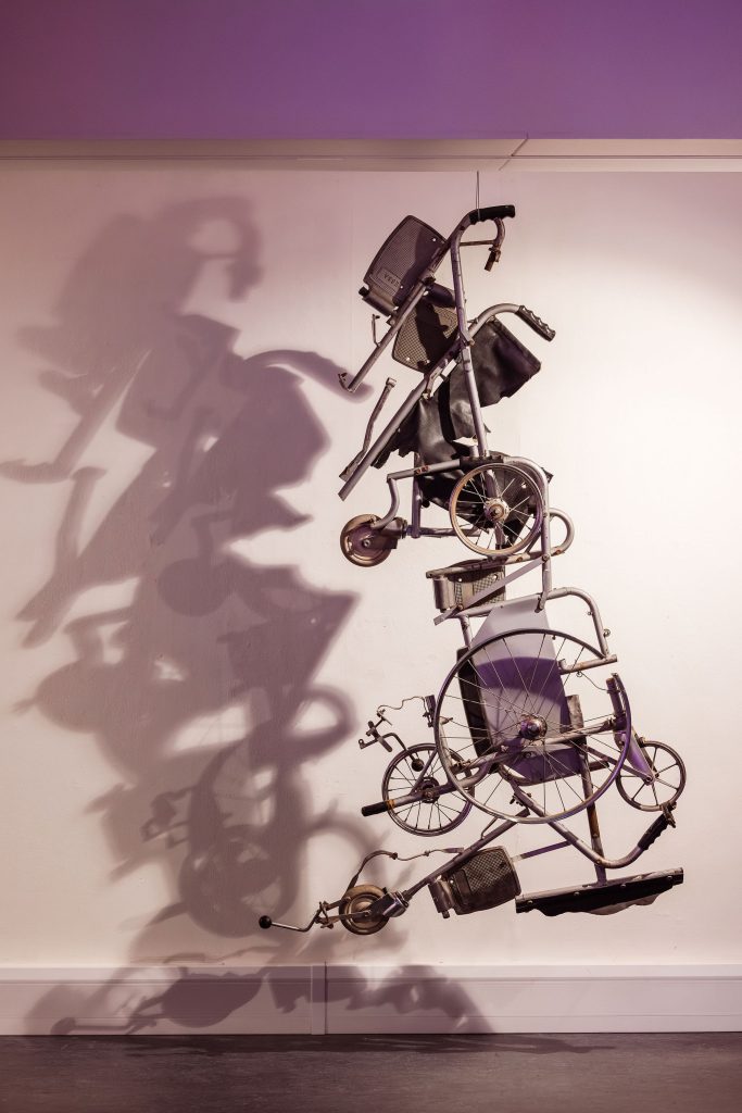 A sculpture of a dismantled metal wheelchair hangs from the ceiling like a mobile