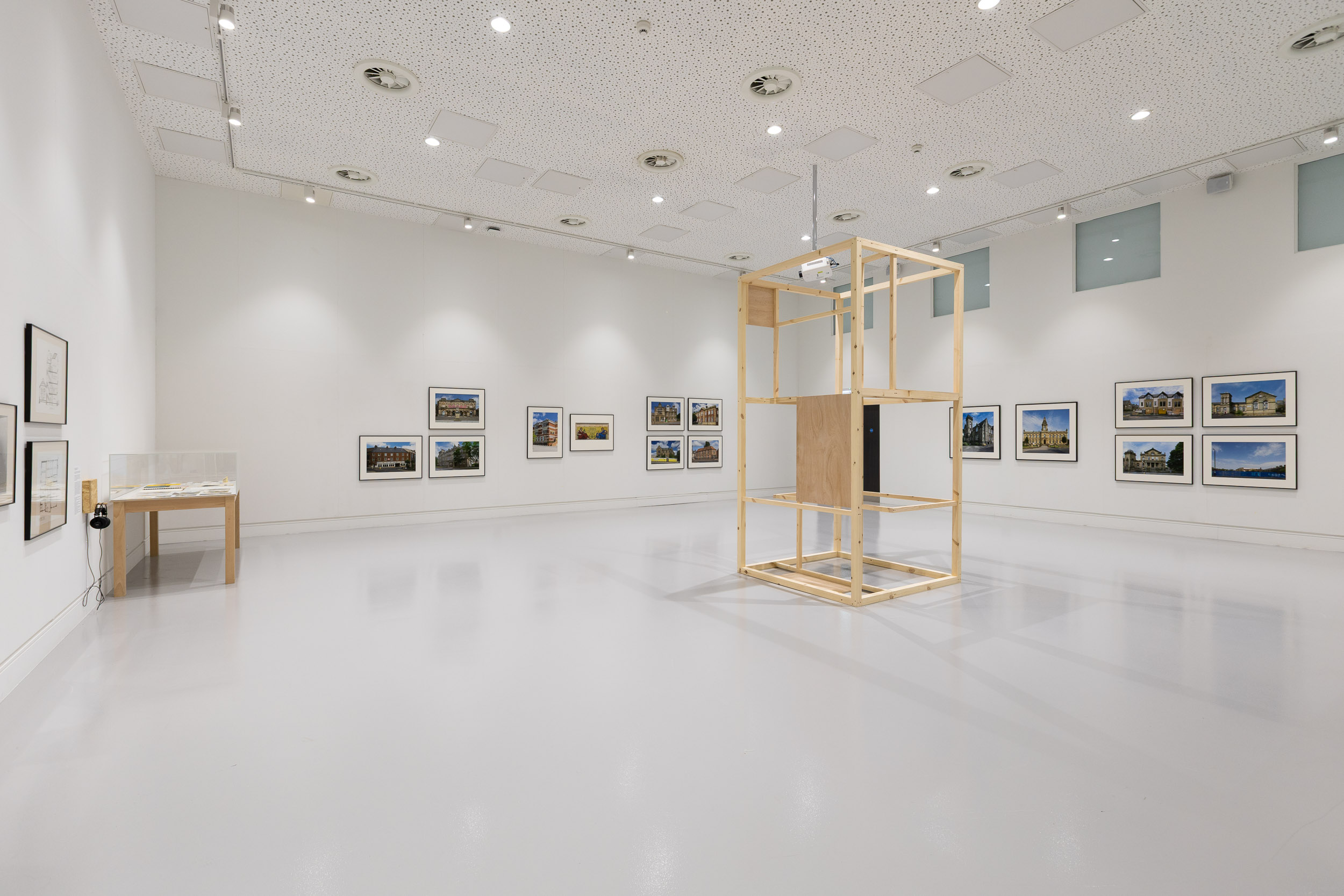 A very bright, clean gallery space. Groupings of two, three and four framed large format photos are on the walls. In the middle of the space a wooden box-like structure and over to the right, a table or vitrine with a pair of headphones hooked on the edge.
