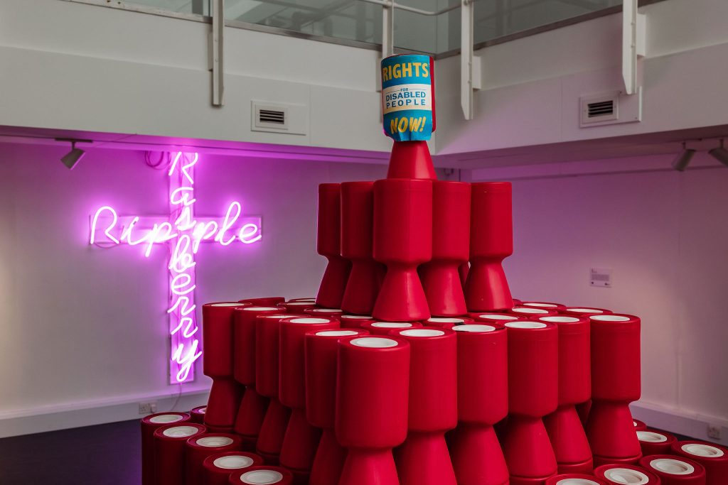 A stack of red charity collection pots in the foreground and in the background an illuminated pink neon crucifix