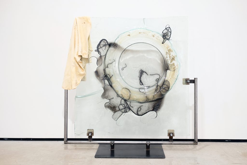 A pane of glass with a circle fused into its surface, surrounded by black curling lines and patches of grey darker glass, supported by metal poles with a soft cream cloth hung on its top left corner